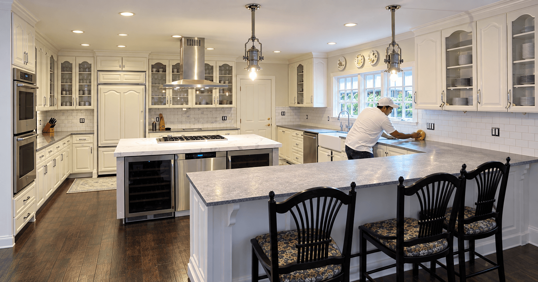 Finished kitchen with a long stone-topped island, white cabinetry, and a person wiping down the countertop